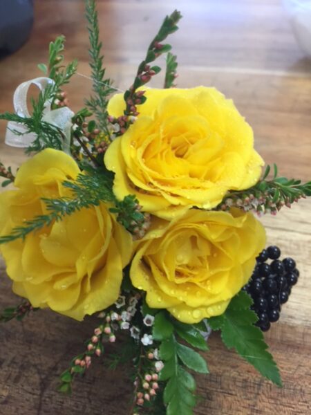 A cluster of three bright yellow roses with water droplets, accented by green fern-like foliage, small white and pink wax flowers, and a cluster of dark purple berries.