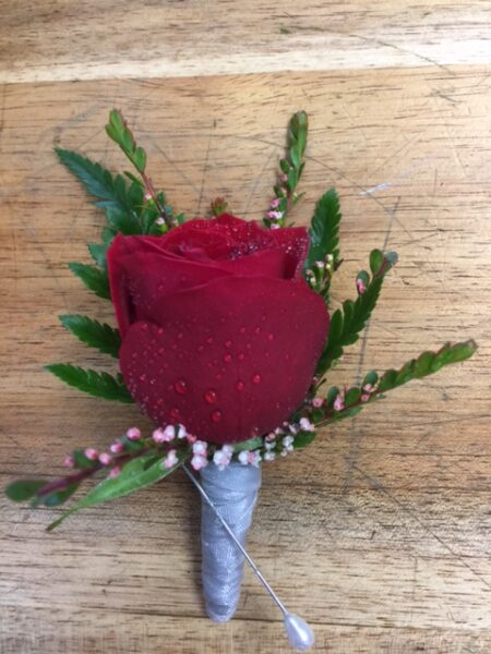 A red rose boutonniere with water droplets, featuring green fern-like foliage and small pink and white wax flowers, with a silver-wrapped stem and a pearl-headed pin.